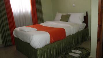 Cama bem arrumada com roupa de cama branca e laranja, cabeceira estofada verde e tapete verde fofo com chinelos.