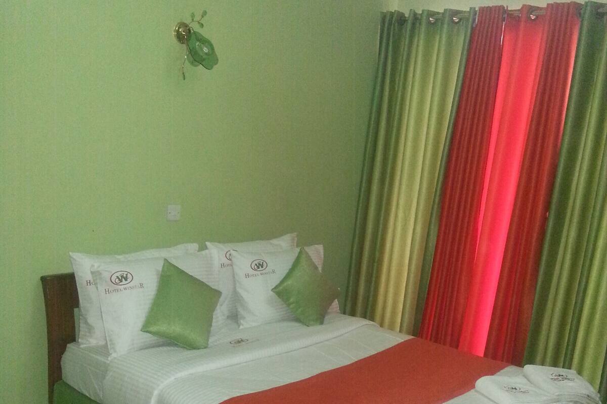 Una cama bien hecha con una colcha roja, almohadas verdes y una base de cama con falda verde. Hay pantuflas blancas sobre una alfombra verde al lado de la cama.