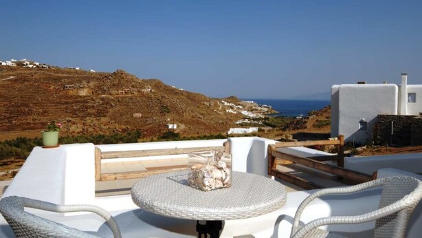 Una terraza con mobiliario blanco de mimbre, una mesa redonda con un centro decorativo, y vistas a un paisaje costero con edificios blancos y el mar.