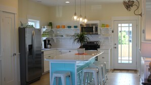 A modern kitchen with a stainless steel refrigerator, a microwave, and a sink. There are three white bar stools and a potted plant on the island.