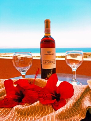A bottle of Granducato Il Santo wine, two wine glasses, and red flowers on a table with a view of the sea. - Asteras Apartments (Sitia)