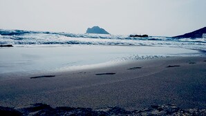 Una spiaggia con una grande formazione rocciosa all'orizzonte, una battigia sabbiosa e impronte che conducono verso l'acqua.