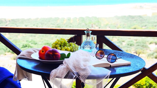 Un tavolo con frutta, una bottiglia, bicchieri e un libro su un balcone con vista sul paesaggio costiero.