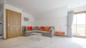 Ein Wohnzimmer mit einem Ecksofa, einem Glastisch, einer Holztür und einem Fenster mit Vorhängen.