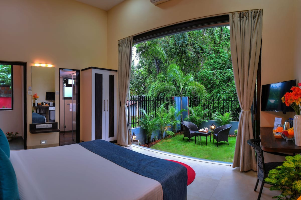 A hotel room with a large bed, a TV, a desk with a vase of flowers, and a view of a lush garden.