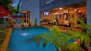 Una piscina interior con pared de piedra y plantas en macetas, zona de bar con asientos y una pantalla de televisión.