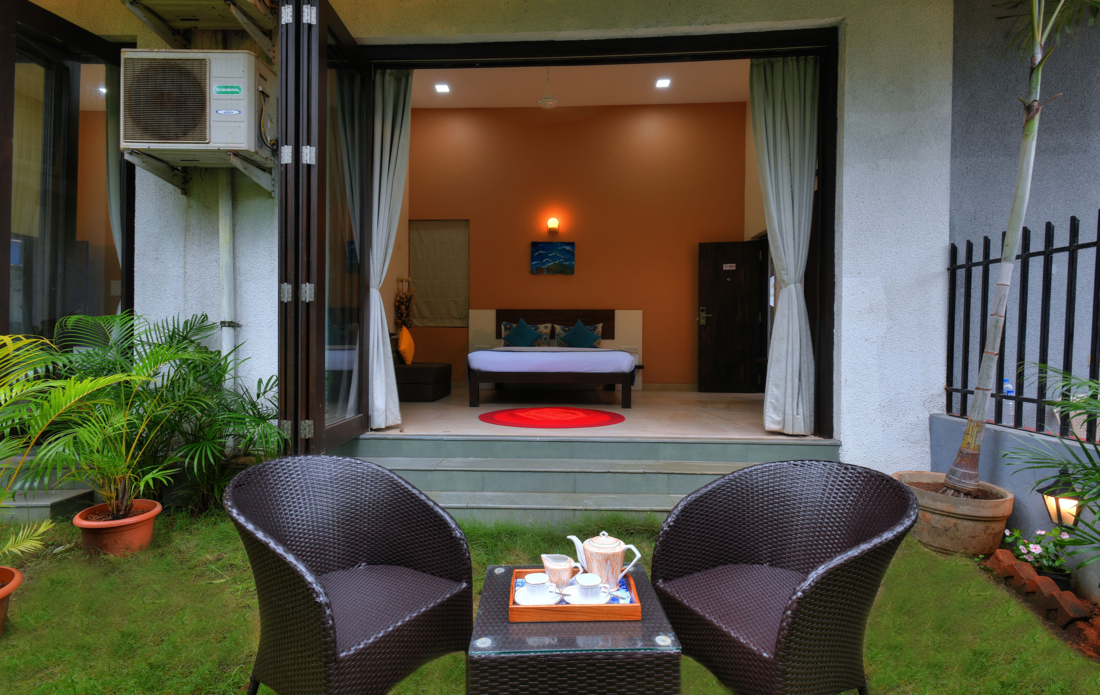 Un dormitorio sin techo con cama, cortinas y una luz nocturna. Hay dos sillones de mimbre y una mesa con una bandeja de té afuera.