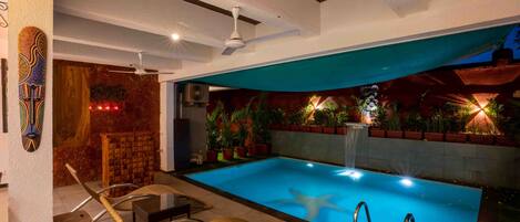An indoor swimming pool with lounge chairs, a ceiling fan, and potted plants.