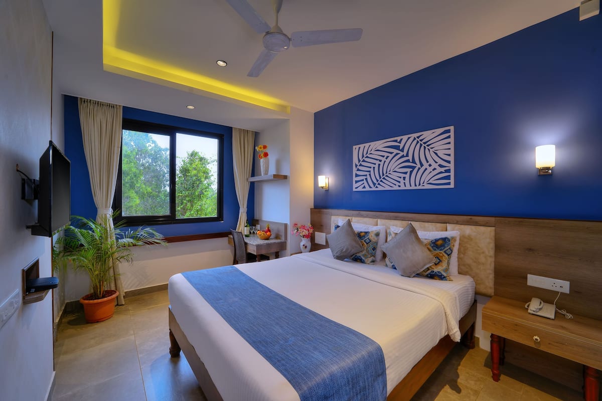 A modern hotel room with a large bed, a ceiling fan, a desk with a chair, a potted plant, and a view of the outdoors.
