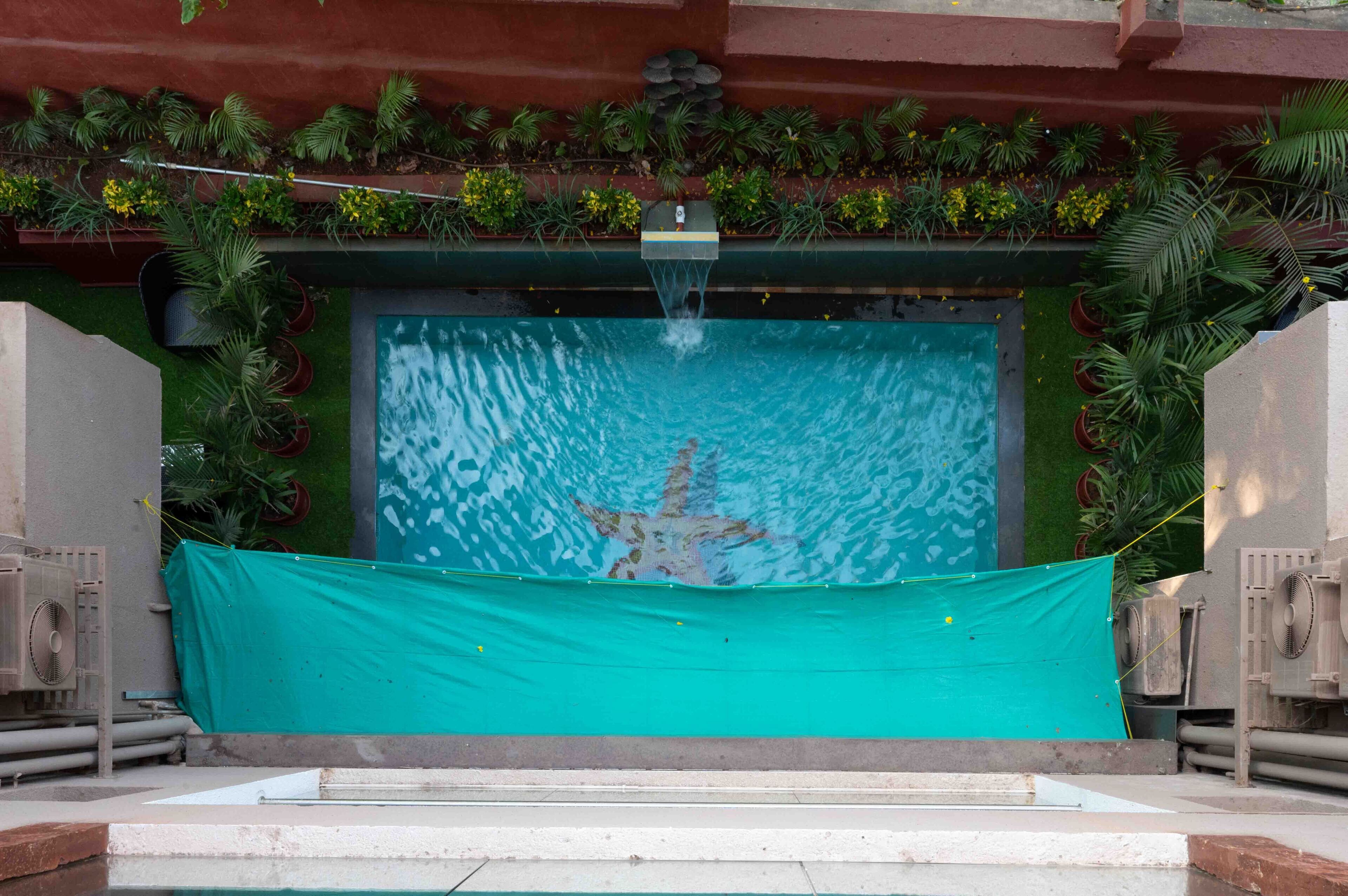 A swimming pool with a waterfall feature, surrounded by potted plants and a green tarp covering part of the pool area.