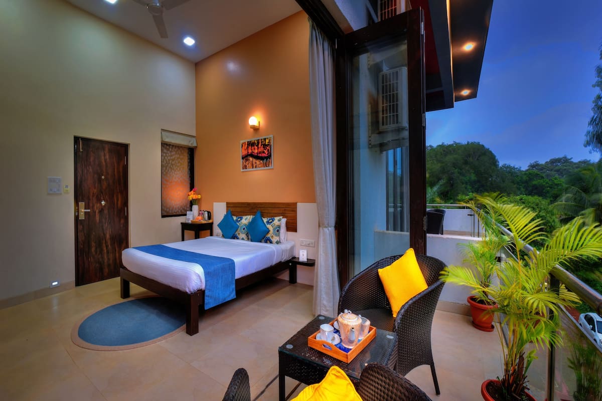 A hotel room with a bed, a nightstand, a table with a tray, a chair, and a balcony with plants.