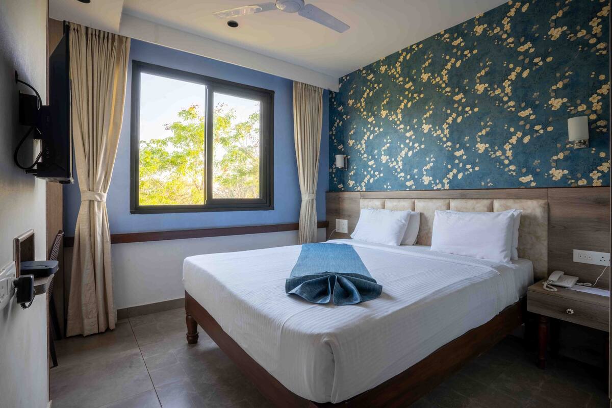 A hotel room with a large bed, a window with curtains, a wall-mounted TV, a bedside table with a phone, and a floral-patterned wall.