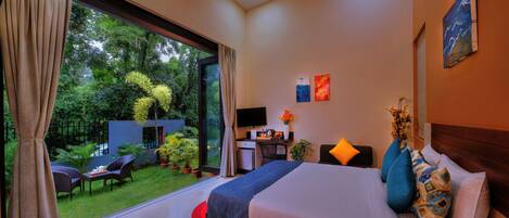 A hotel room with a large bed, a desk with a computer, a TV, and a view of a garden.