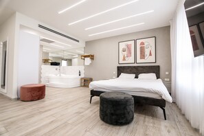 A modern bedroom with a large bed, a round ottoman, a wooden bench, a white bathtub, and a large mirror.