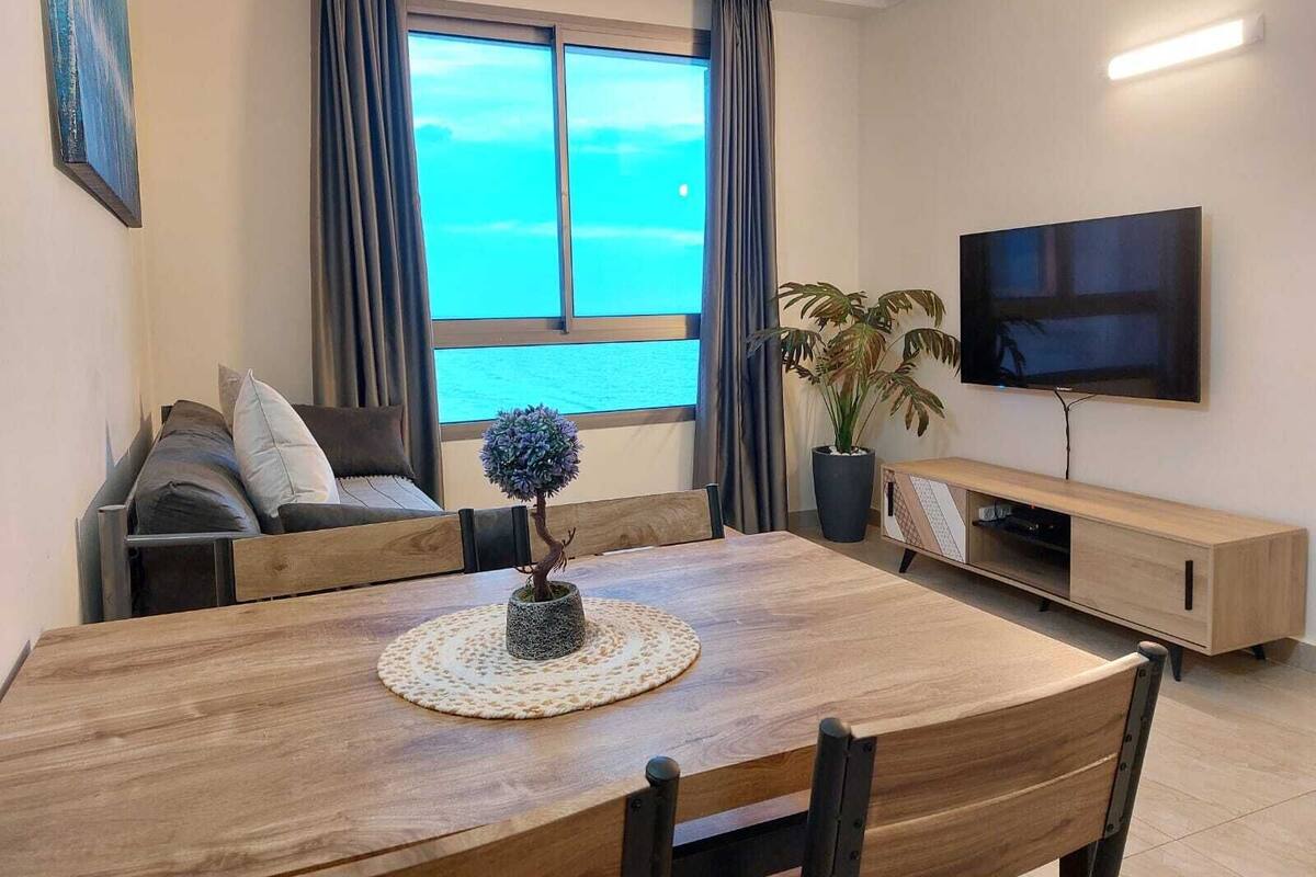 Un salon moderne avec une table à manger en bois, un canapé, un téléviseur et une vue sur la mer grâce à de grandes fenêtres.
