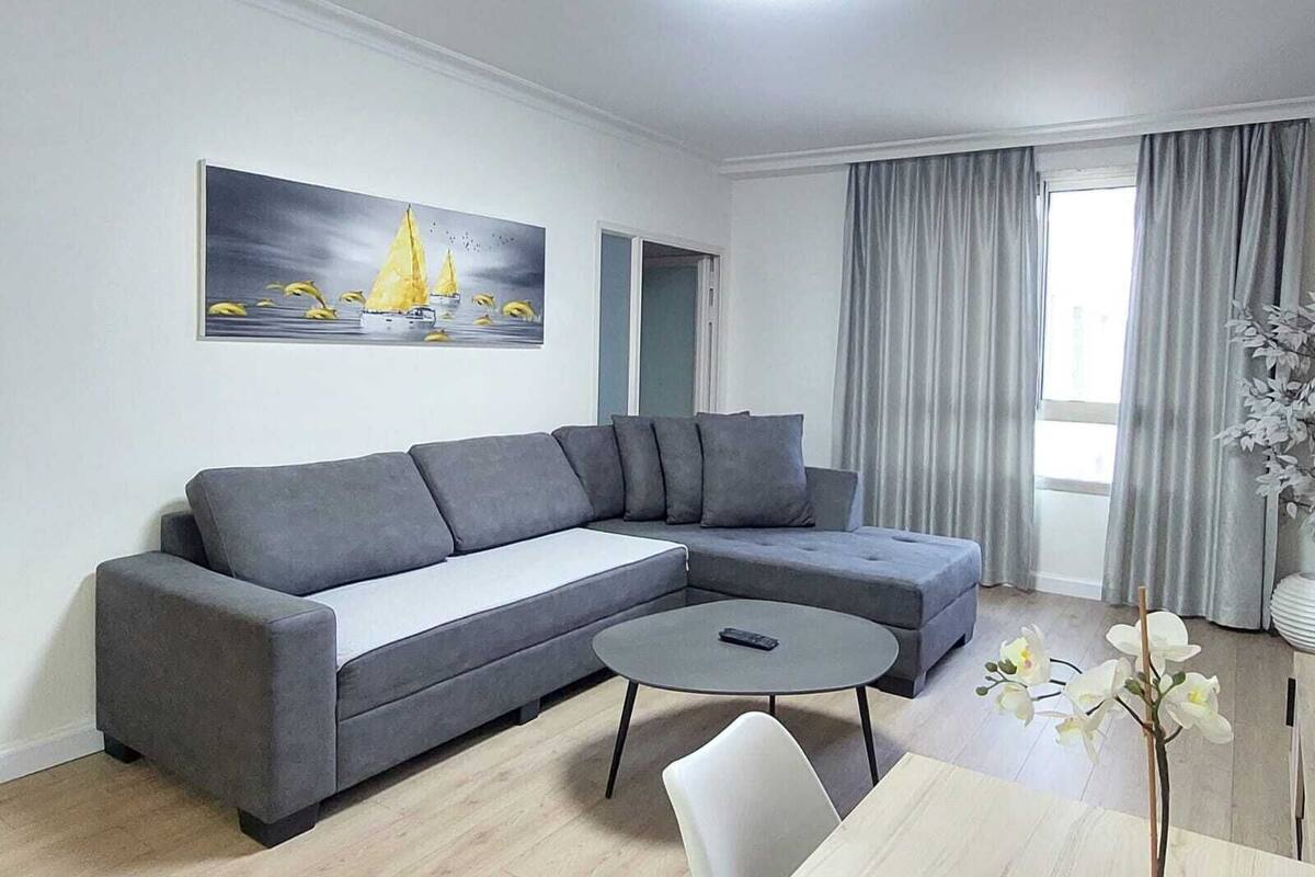 Un salon moderne avec un canapé d’angle gris, une table basse ronde et un tableau représentant un voilier accroché au mur.