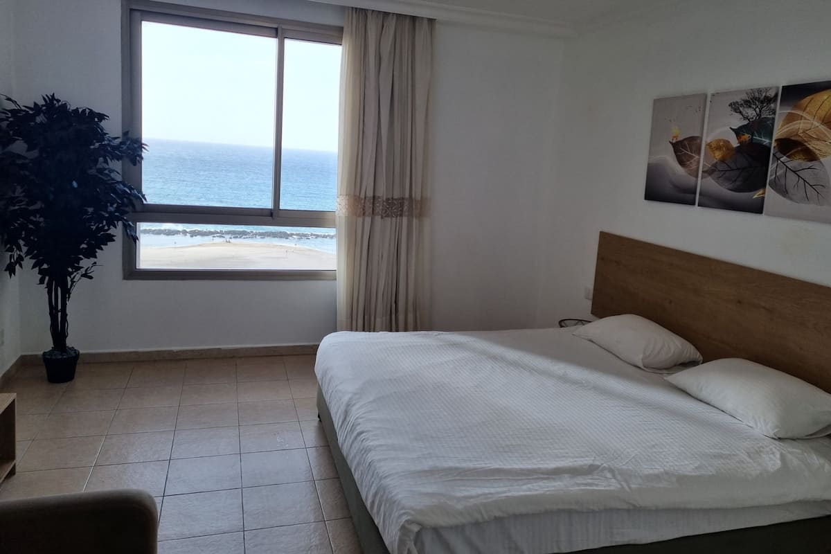 Une chambre d’hôtel avec un lit, une fenêtre donnant sur la plage, une plante en pot et un mur décoré d’œuvres d’art.