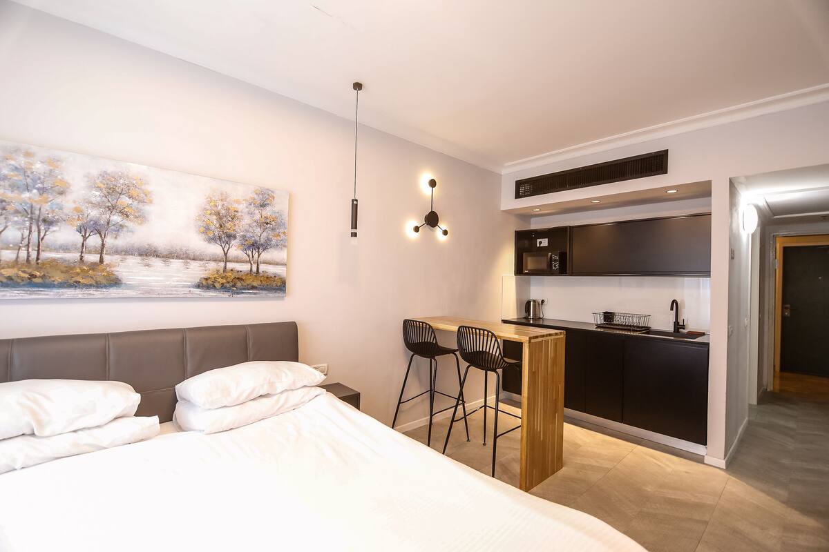 Une chambre d’hôtel moderne comprenant un lit, un bureau avec deux chaises, un coin cuisine et un tableau au mur.