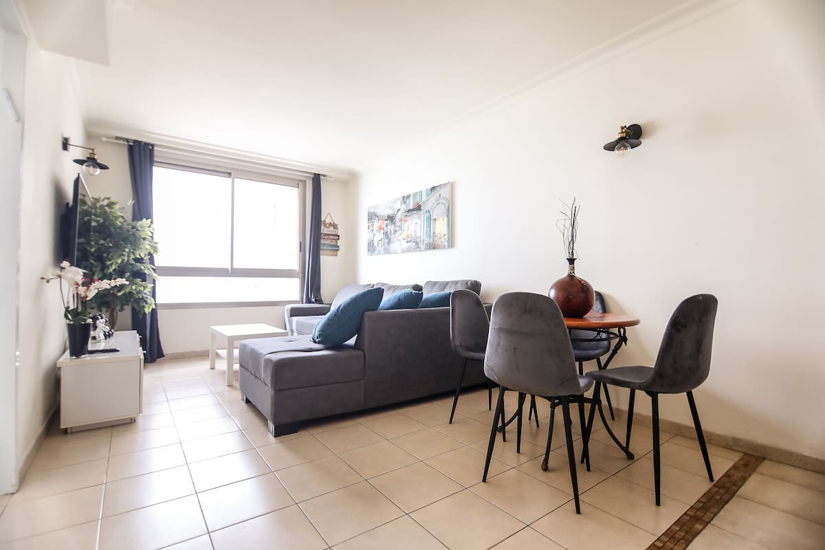 Un salon moderne avec un canapé gris, une table à manger avec des chaises, une plante en pot et une grande fenêtre.
