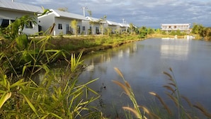A row of modern, single-story buildings with large windows, situated beside a calm river. - Coffee&Homestay568 Branch 2 (Muang Suang)