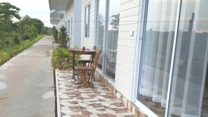 Un espacio al aire libre techado con piso de baldosas, una mesa de madera y sillas, junto a un edificio con ventanas grandes y fachada blanca.
