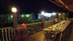 Una larga terraza de madera con mesas dispuestas para comer, rodeada por una barandilla de madera e iluminada por luces esféricas.