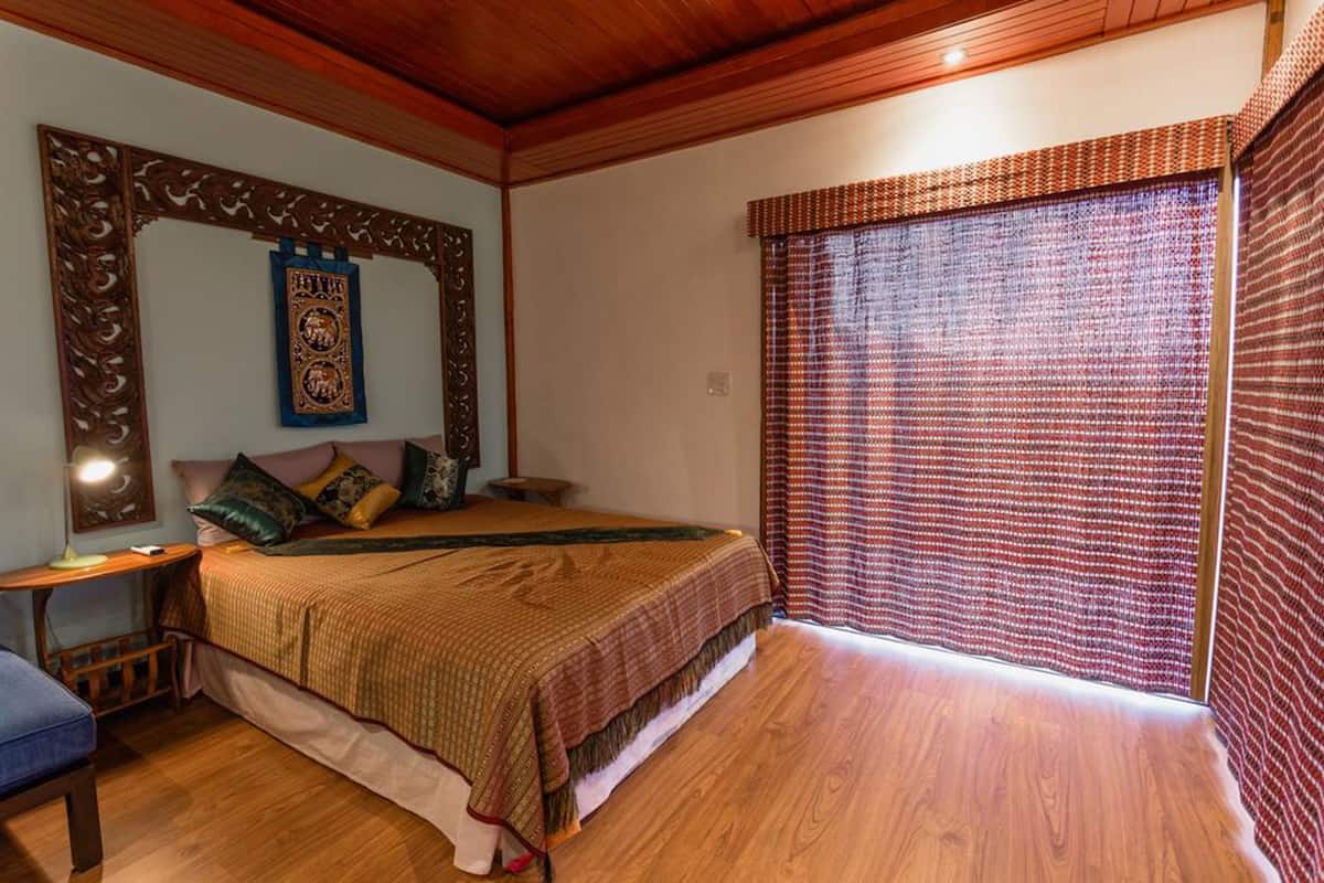 A bedroom with a wooden bed, a large mirror, a bedside table, and a window with patterned curtains.