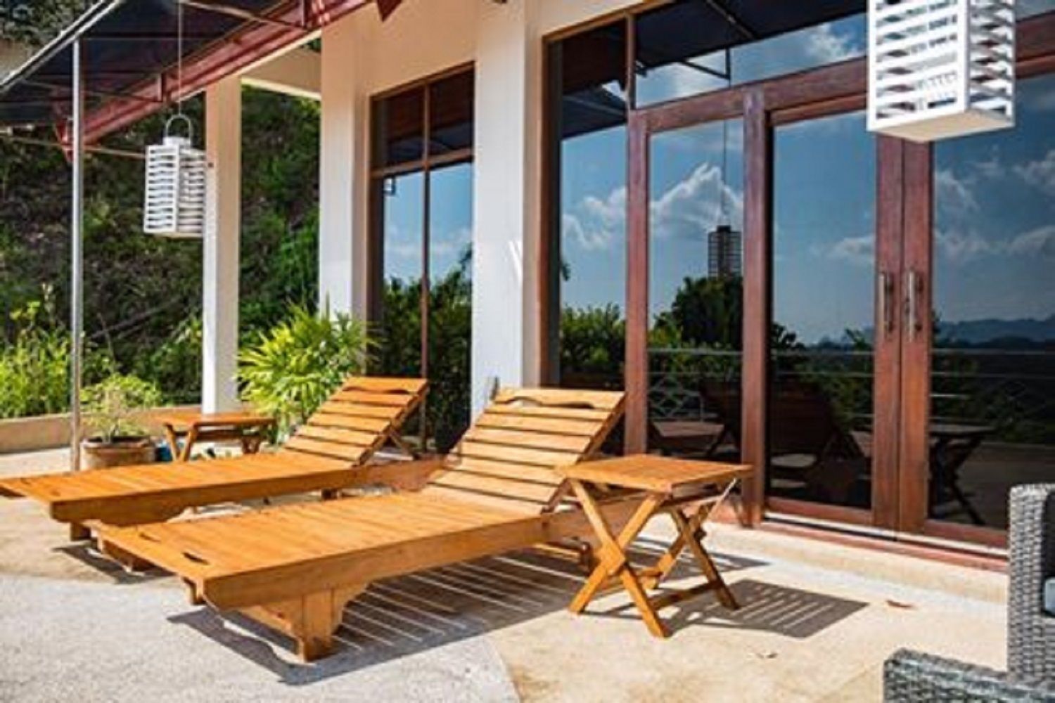 A covered outdoor seating area with wooden lounge chairs and a table, large glass doors, and a view of the landscape.