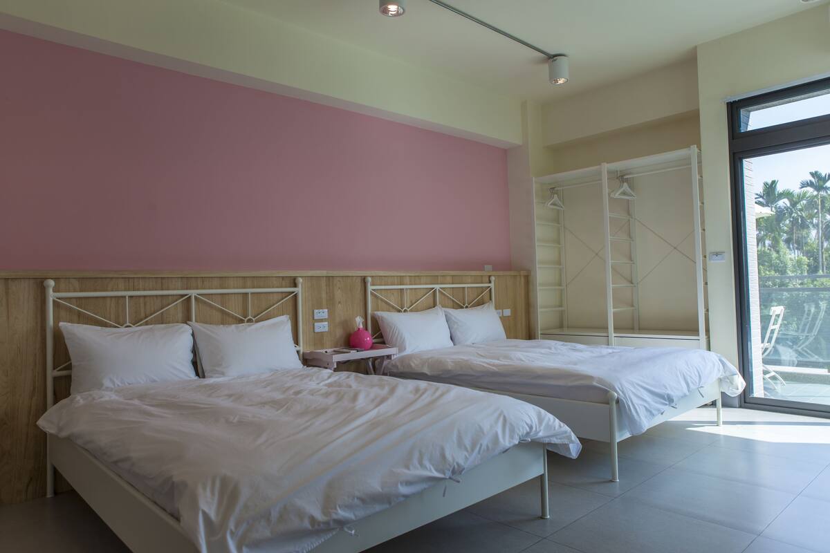 Un dormitorio con dos camas, cabeceros de madera, pared rosa y un amplio ventanal con vistas a palmeras.