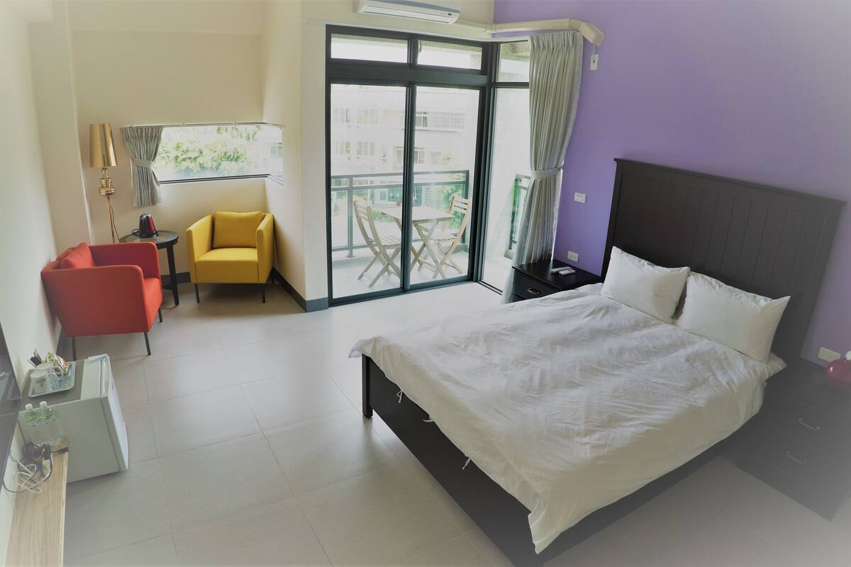 Habitación de hotel con una cama grande, una silla roja, una silla amarilla, una mesita y un balcón con vistas al exterior.