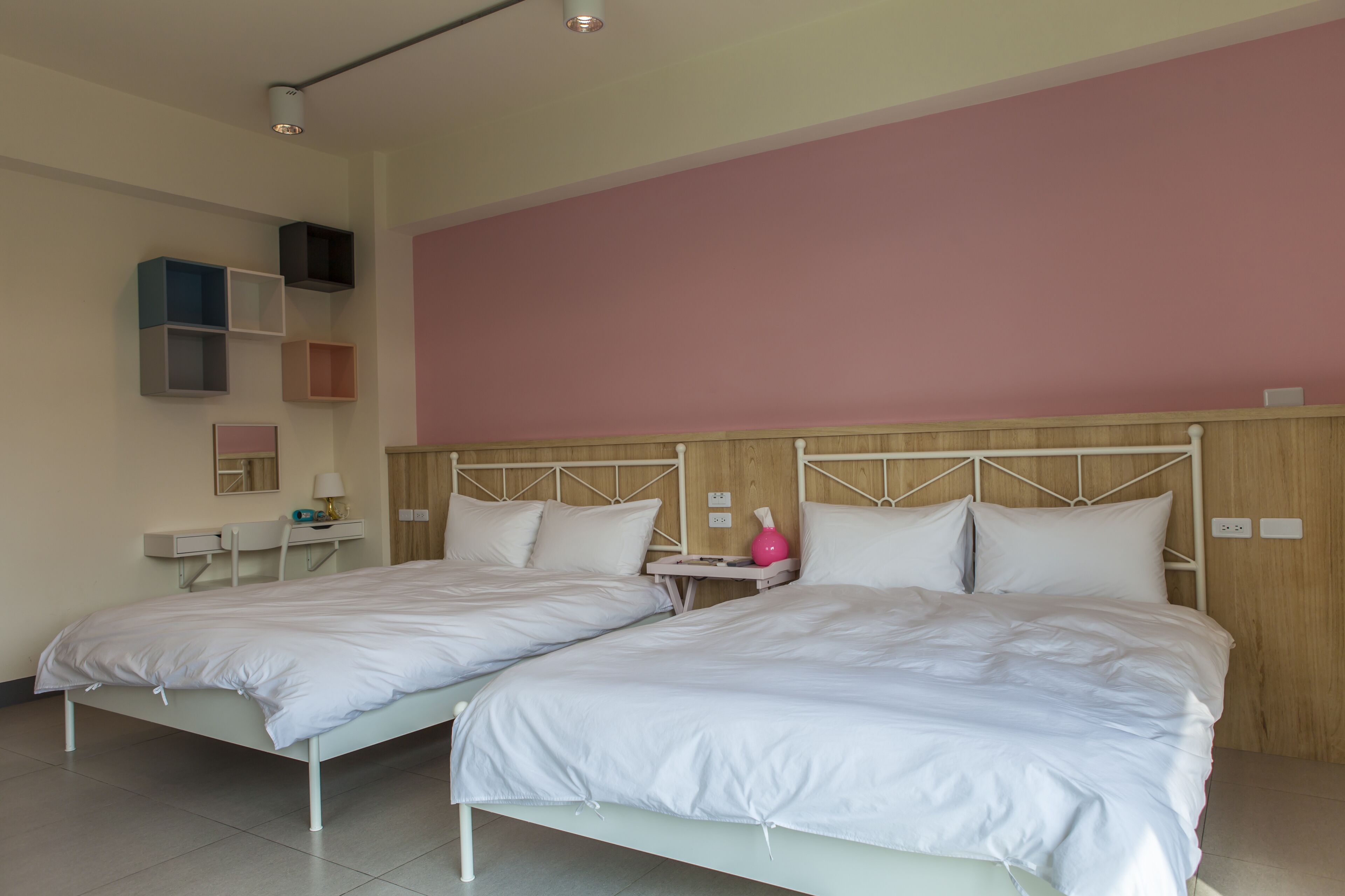 A room with two beds, a bedside table, a lamp, and a shelf with storage boxes.