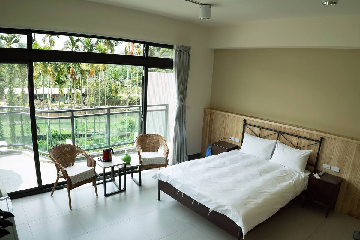 Habitación de hotel con una cama, dos sillas, una mesita, una mesita de noche y vistas a la vegetación a través de grandes ventanales.