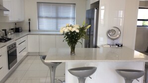 A modern kitchen with white cabinets, a central island with bar stools, a stainless steel refrigerator, and a window with blinds. - 43 Ocean View Drive (Cape Town)