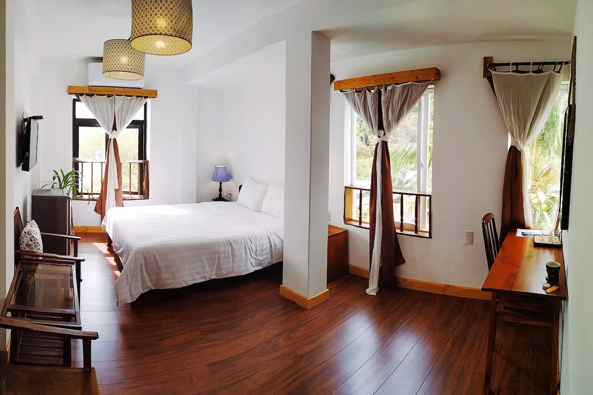 A spacious bedroom with a large bed, wooden flooring, a desk, and a chair. There are windows with curtains, a hanging light fixture, and a wall-mounted TV.