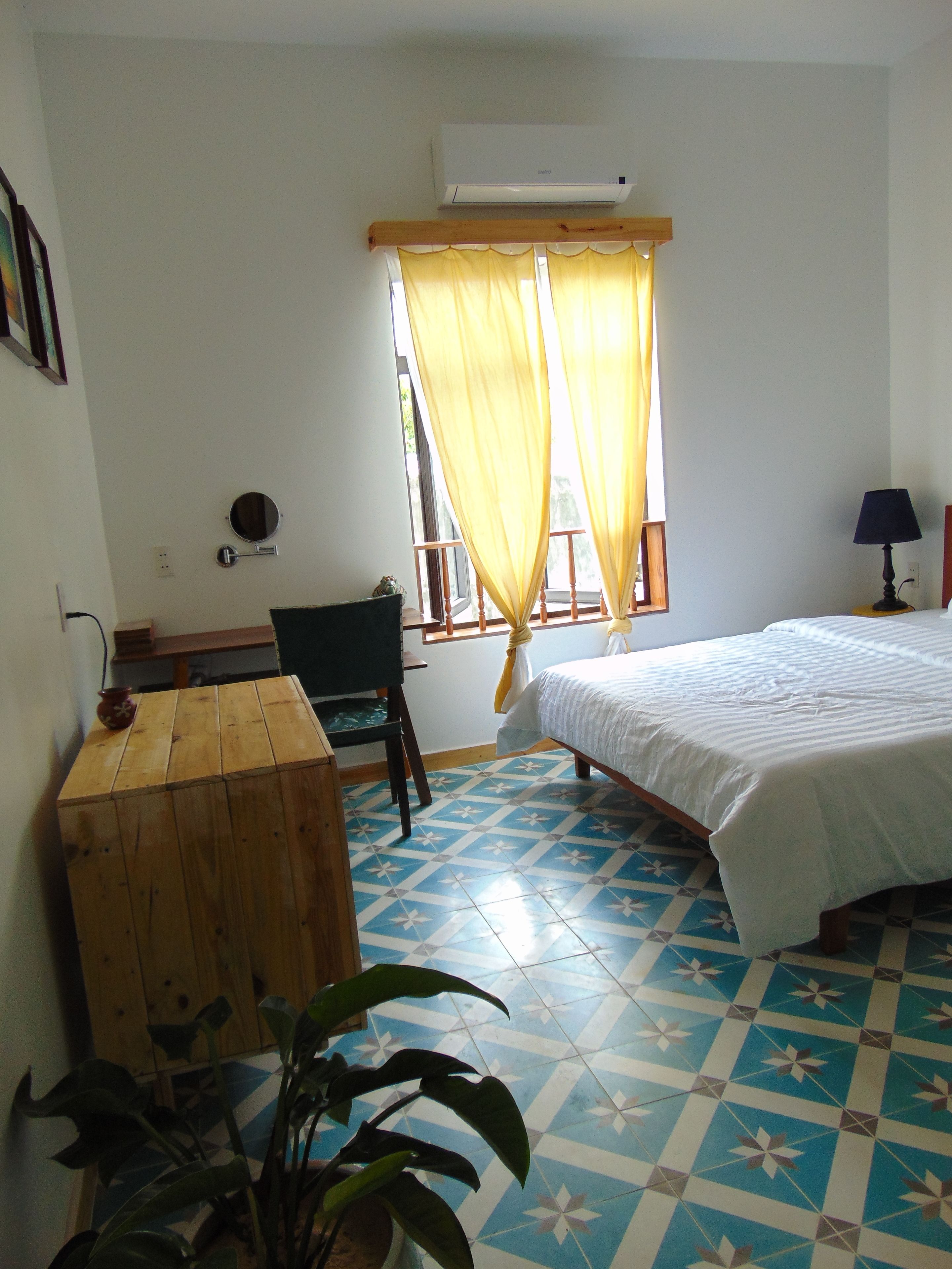 A room with a bed, a desk, a chair, a potted plant, and a window with yellow curtains.