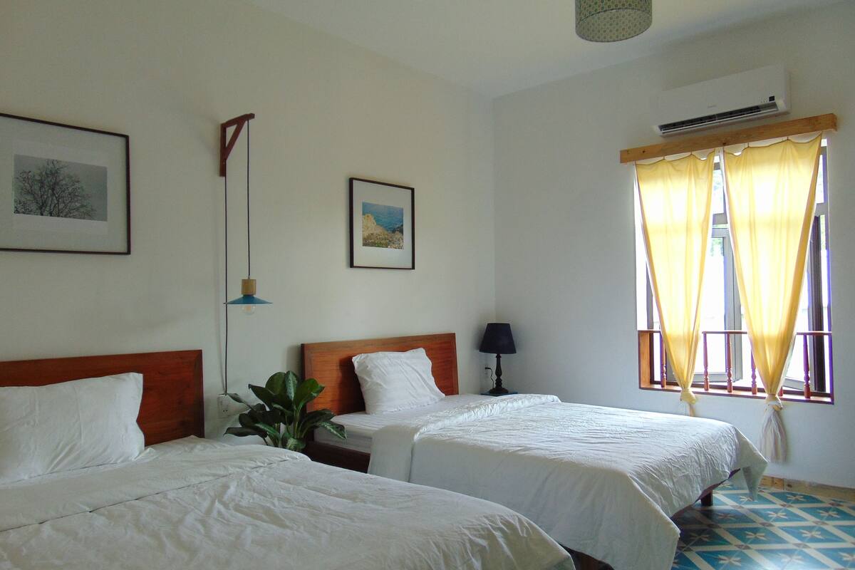 A hotel room with two beds, a nightstand, a lamp, a potted plant, and a window with yellow curtains.