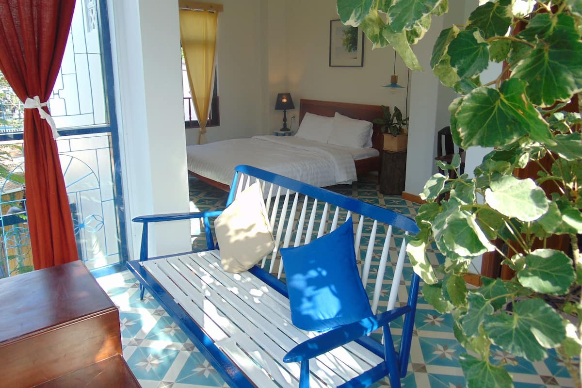 A room with a bed, a window with curtains, a plant, and a bench.