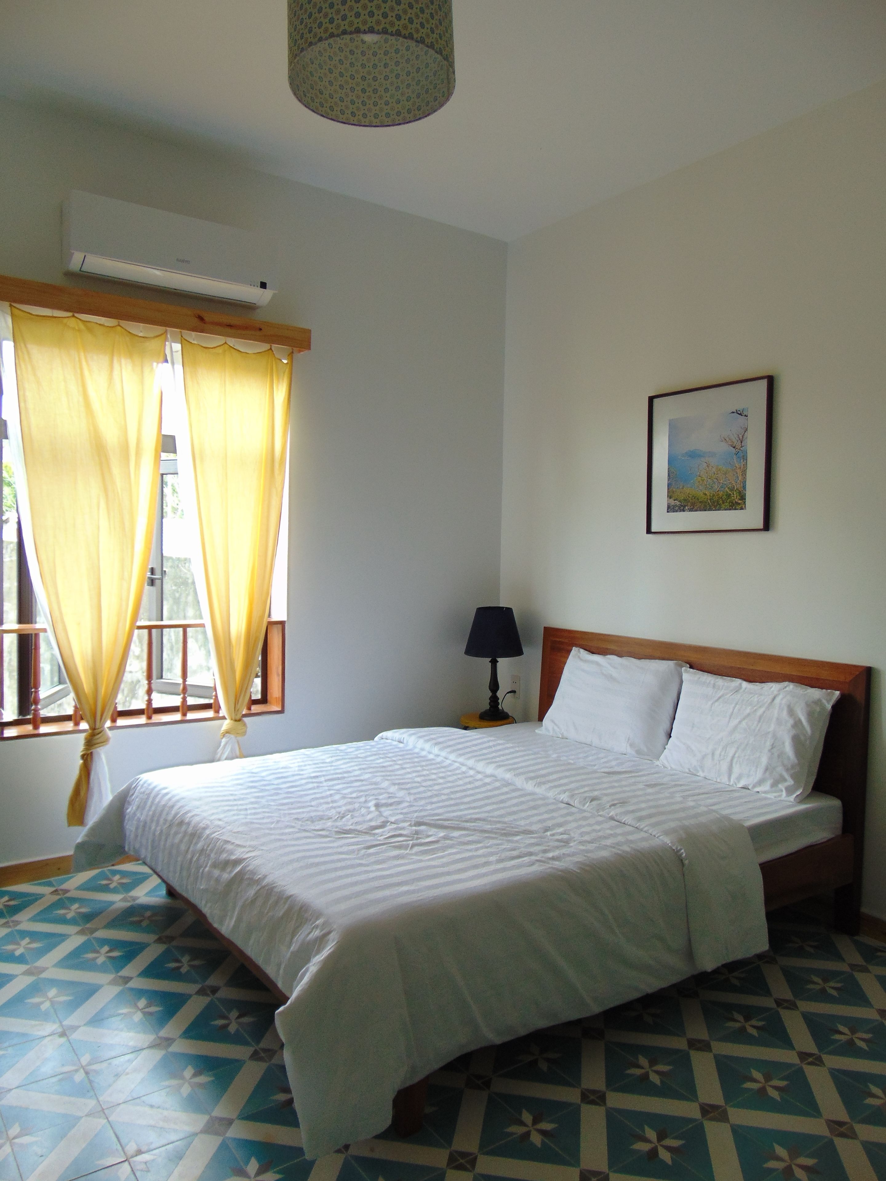 A bedroom with a bed, a nightstand, a lamp, a window with curtains, a picture on the wall, and a patterned floor.