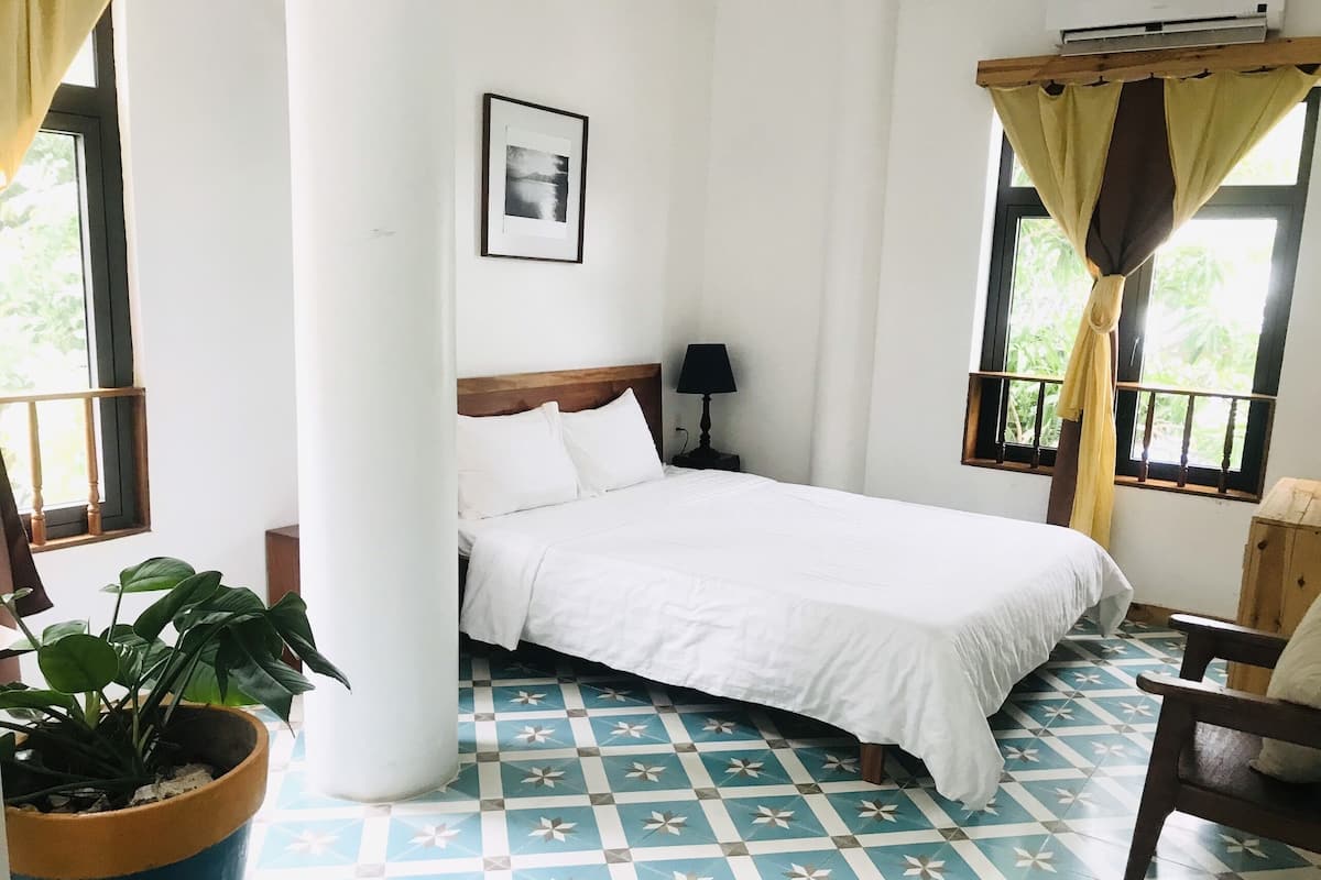 A bedroom with a bed, a nightstand, a potted plant, a window with curtains, and a patterned floor.