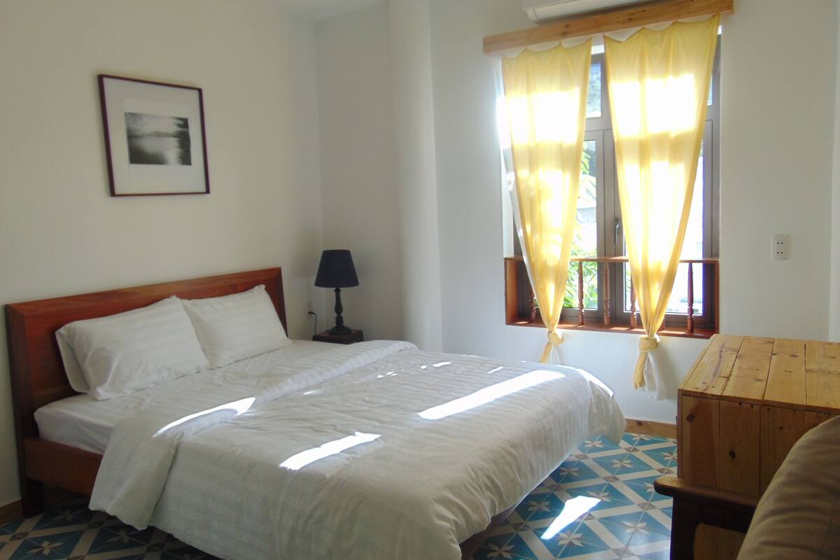 A bedroom with a bed, a bedside table with a lamp, a window with curtains, and a picture on the wall.