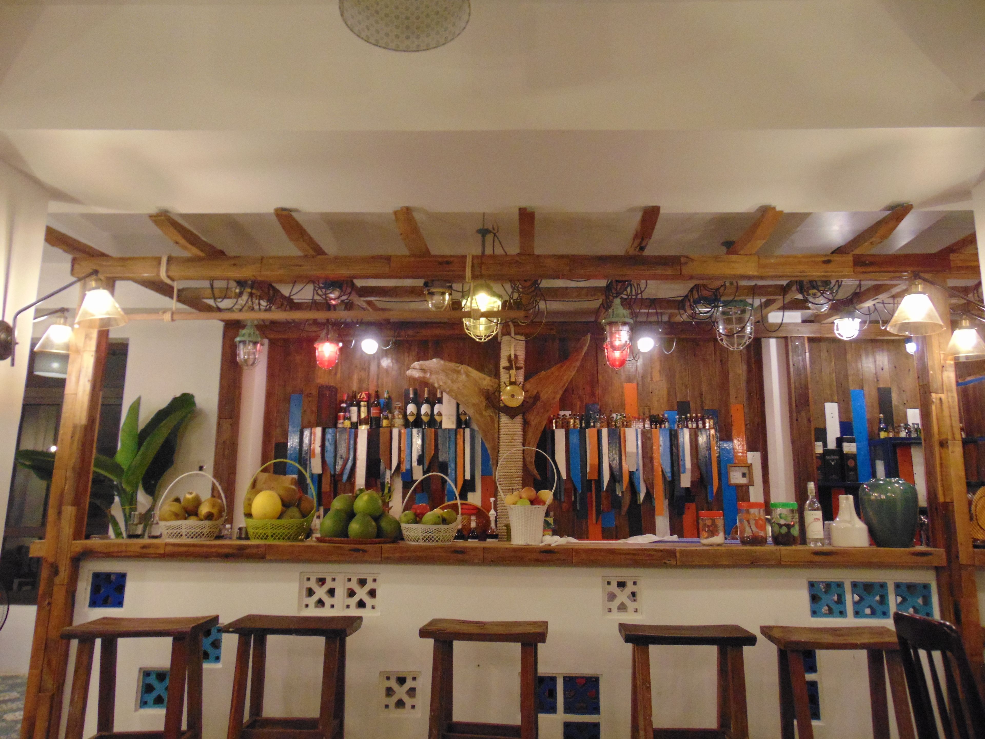 A bar area with wooden beams, hanging lights, and a variety of bottles and fruits displayed on the counter.