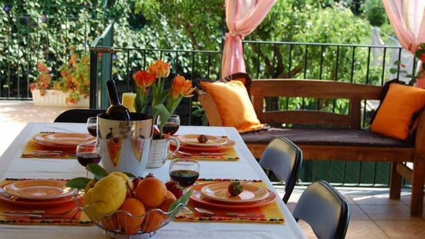 An outdoor dining area with a table set for four, surrounded by greenery and a wooden bench with orange cushions. - House / Villa - Giarre (Giarre)
