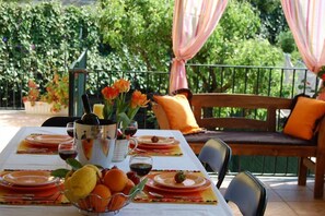 An outdoor dining area with a table set for four, surrounded by greenery and a wooden bench with orange cushions. - House / Villa - Giarre (Giarre)