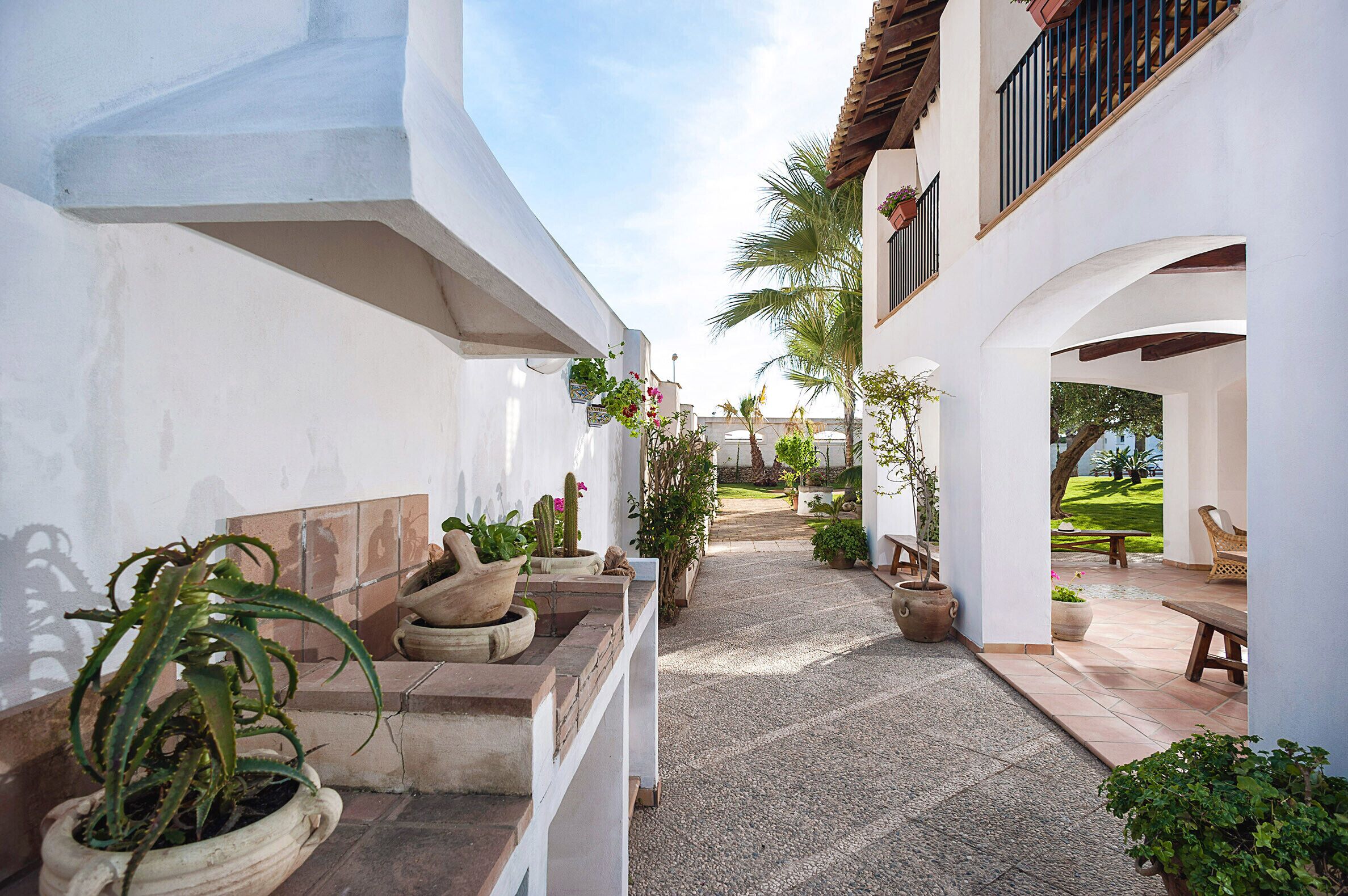 A narrow pathway flanked by white walls and potted plants, leading to an outdoor seating area with a bench and a table.