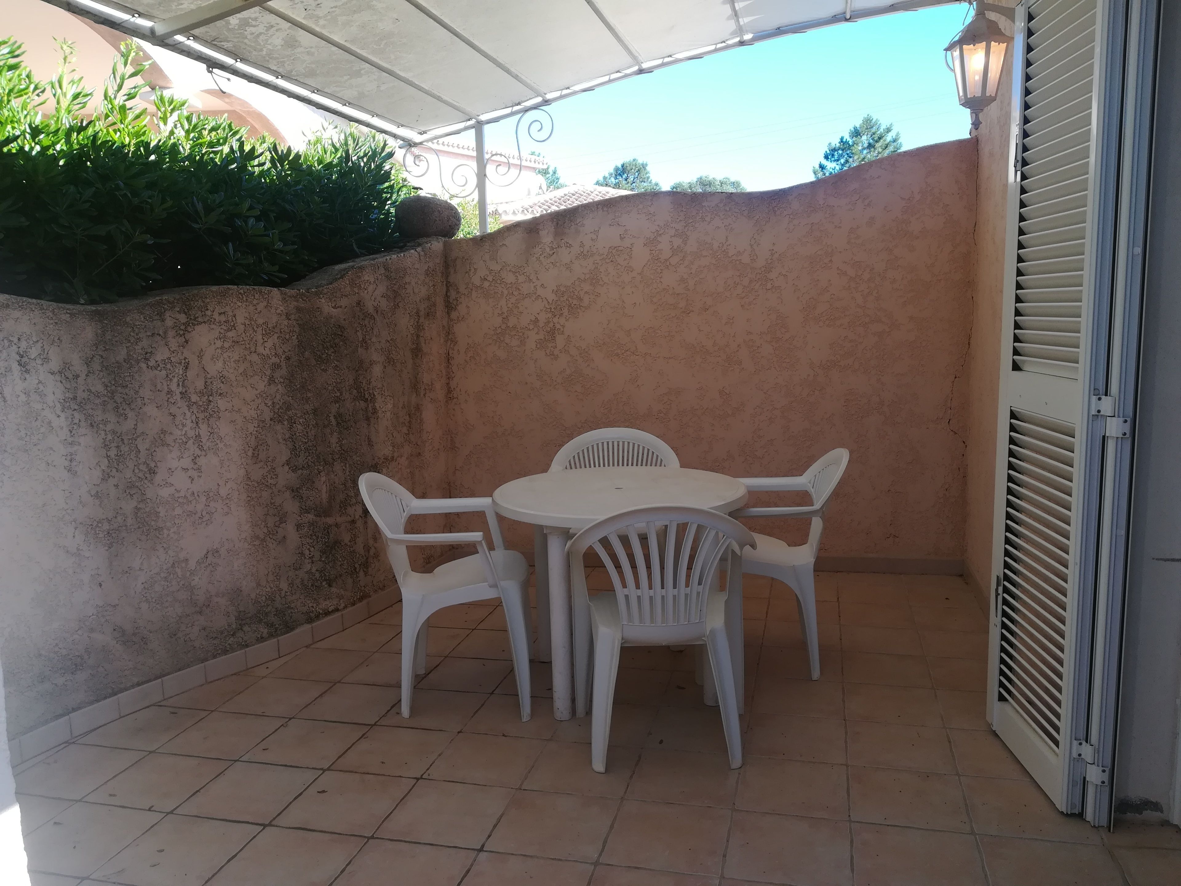 Un patio con una mesa de plástico blanco y cuatro sillas, suelo de baldosas, pared con superficie texturizada y una puerta entreabierta.