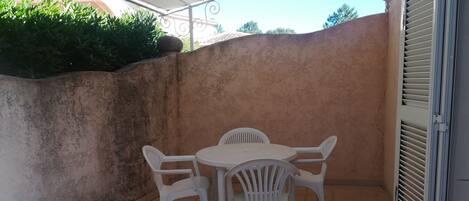 Un patio con una mesa de plástico blanco y cuatro sillas, suelo de baldosas, pared con superficie texturizada y una puerta entreabierta.