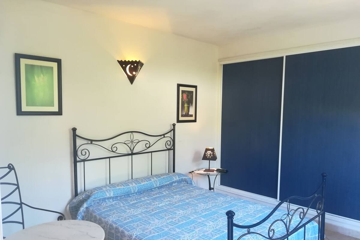 A bedroom with a bed, a small table, a wall-mounted lamp, and a picture frame.
