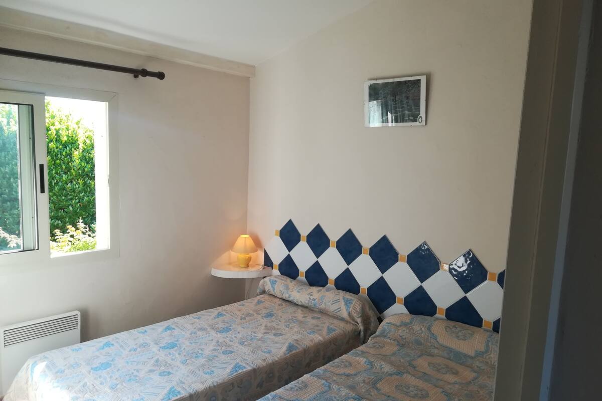 A room with two beds, a small table with a lamp, a window with a view of greenery, and a wall-mounted shelf.