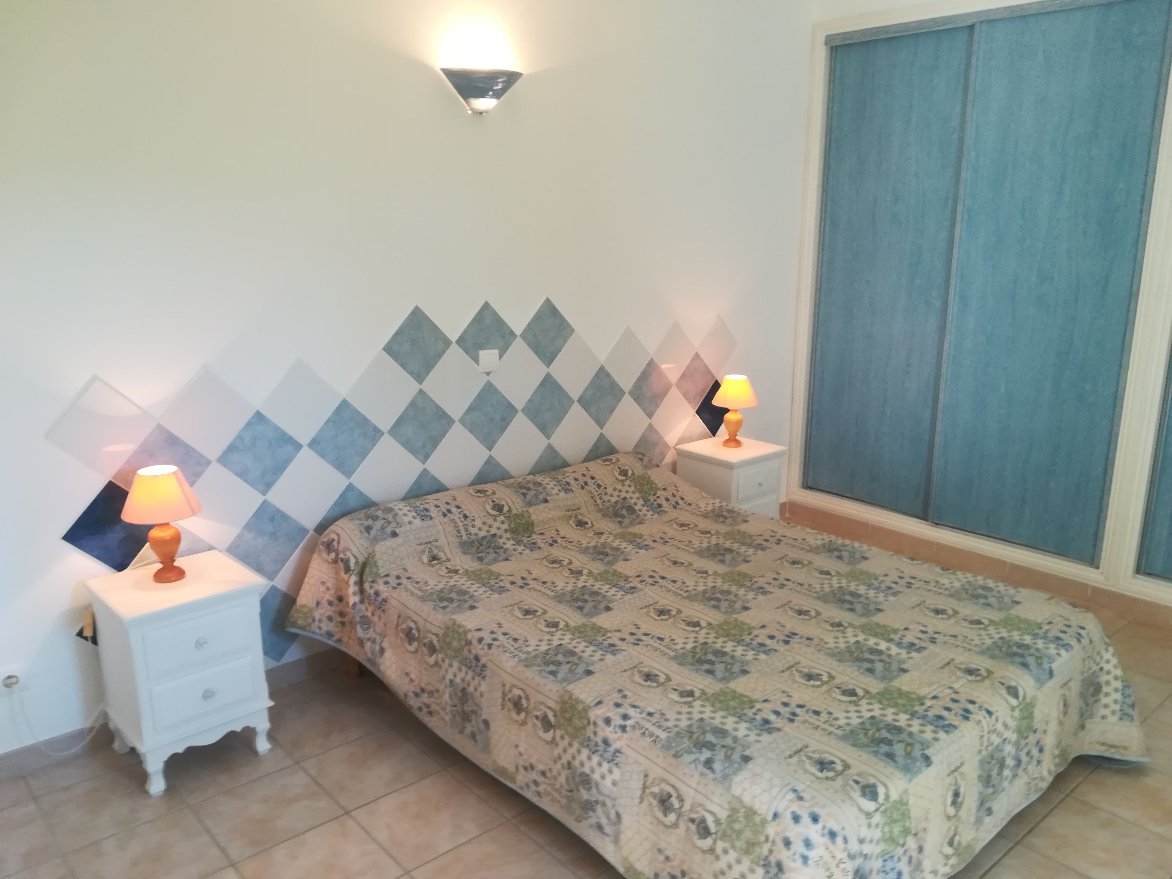 A bedroom with a patterned bedspread, two bedside lamps, a white nightstand, and a wall with a diamond-patterned tile design.