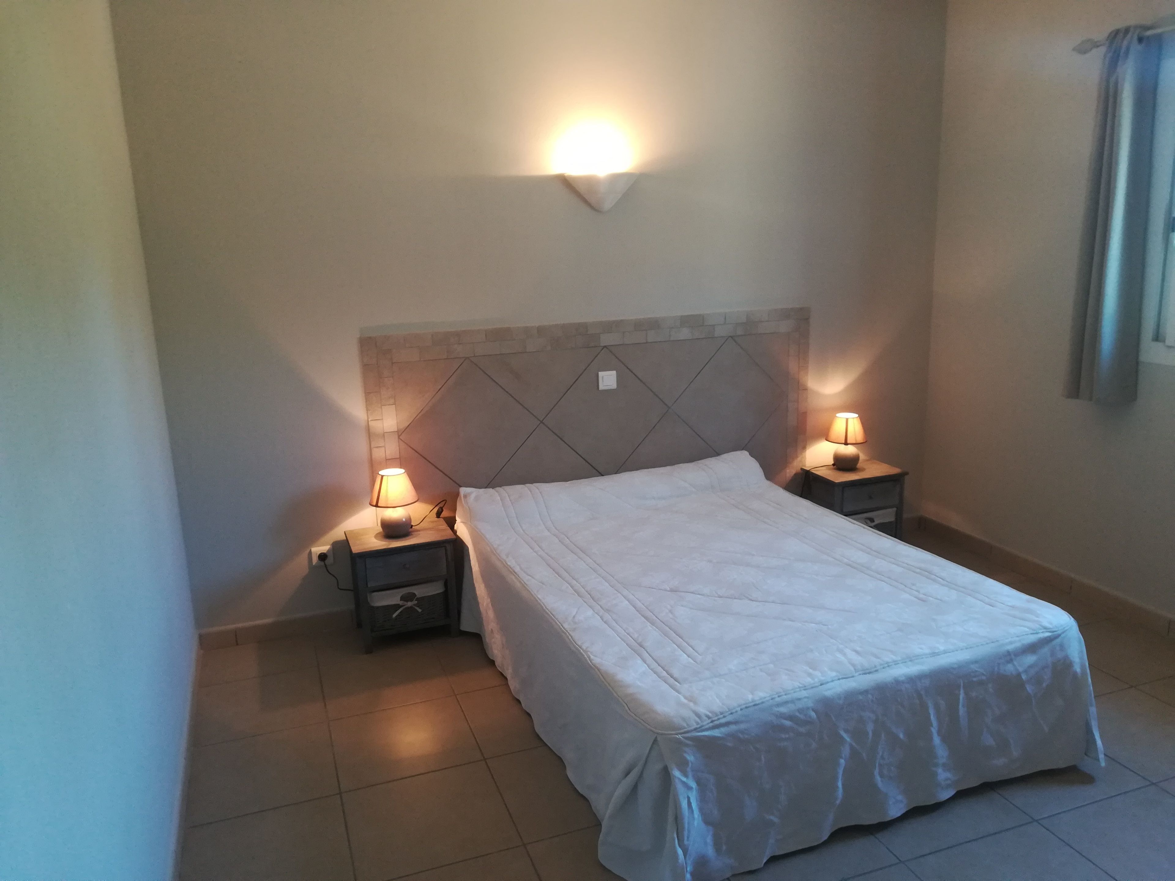 Una cama bien hecha con una colcha blanca y dos lámparas de noche sobre mesitas de madera.